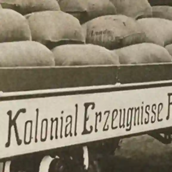 Stilisiertes, altes Foto eines Motivwagens, der mit gefüllten Säcken beladen ist. Man erkennt den darauf stehenden Schriftzug: "Kolonial Erzeugnisse ... ".