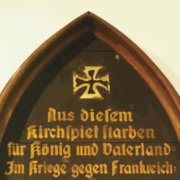 Stilisierter Ausschnitt einer hölzernen, schwarz grundierten Gedenktafel in Form eines Spitzbogens mit goldfarbener Schrift und gemaltem Eisernen Kreuz. Die Transkription findet sich im PDF.